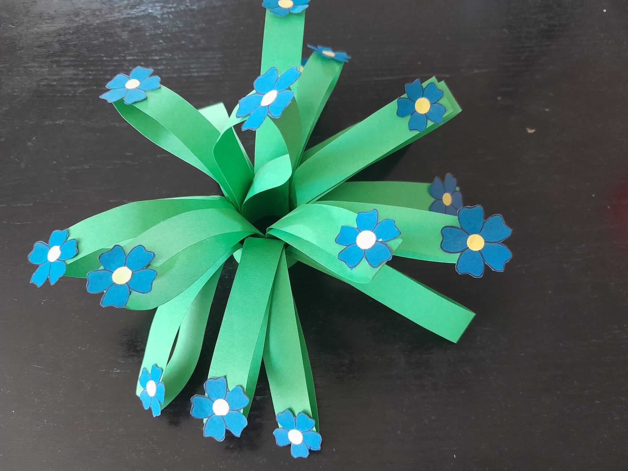 A paper craft featuring green strips arranged like grass with small blue flowers attached to the ends. - Home Instead