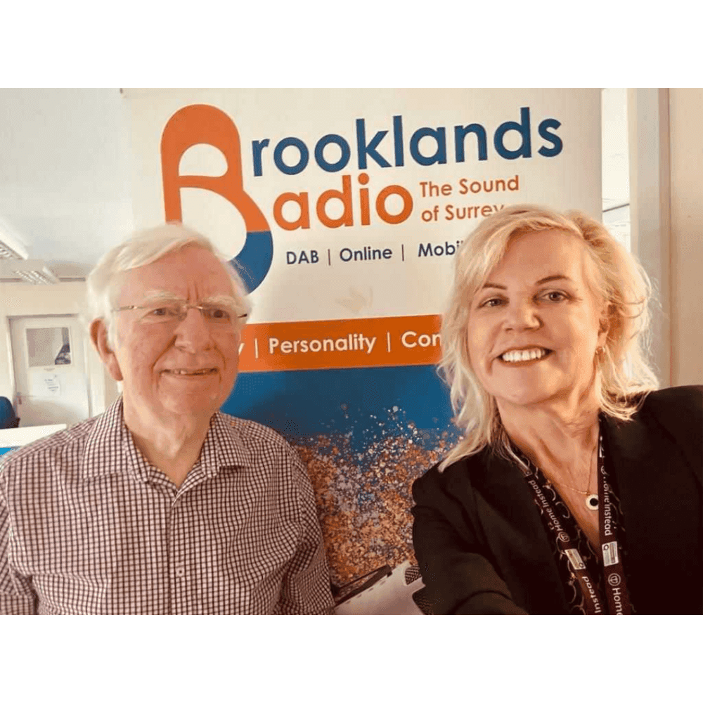 Two people smiling in front of a Brooklands Radio banner that reads "The Sound of Surrey - DAB | Online | Mobile". - Home Instead