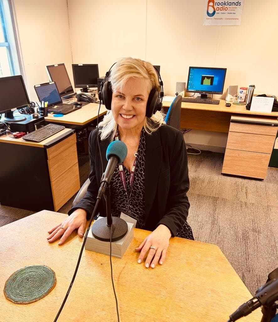 A woman with headphones speaks into a microphone at a radio station studio, surrounded by desks and computers. - Home Instead