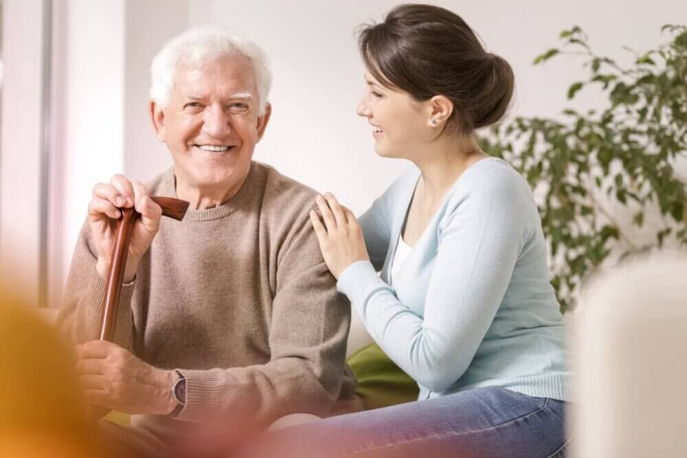 An elderly man holding a cane smiles while a young woman beside him places a hand on his shoulder and smiles back. - Home Instead Bournemouth & Christchurch