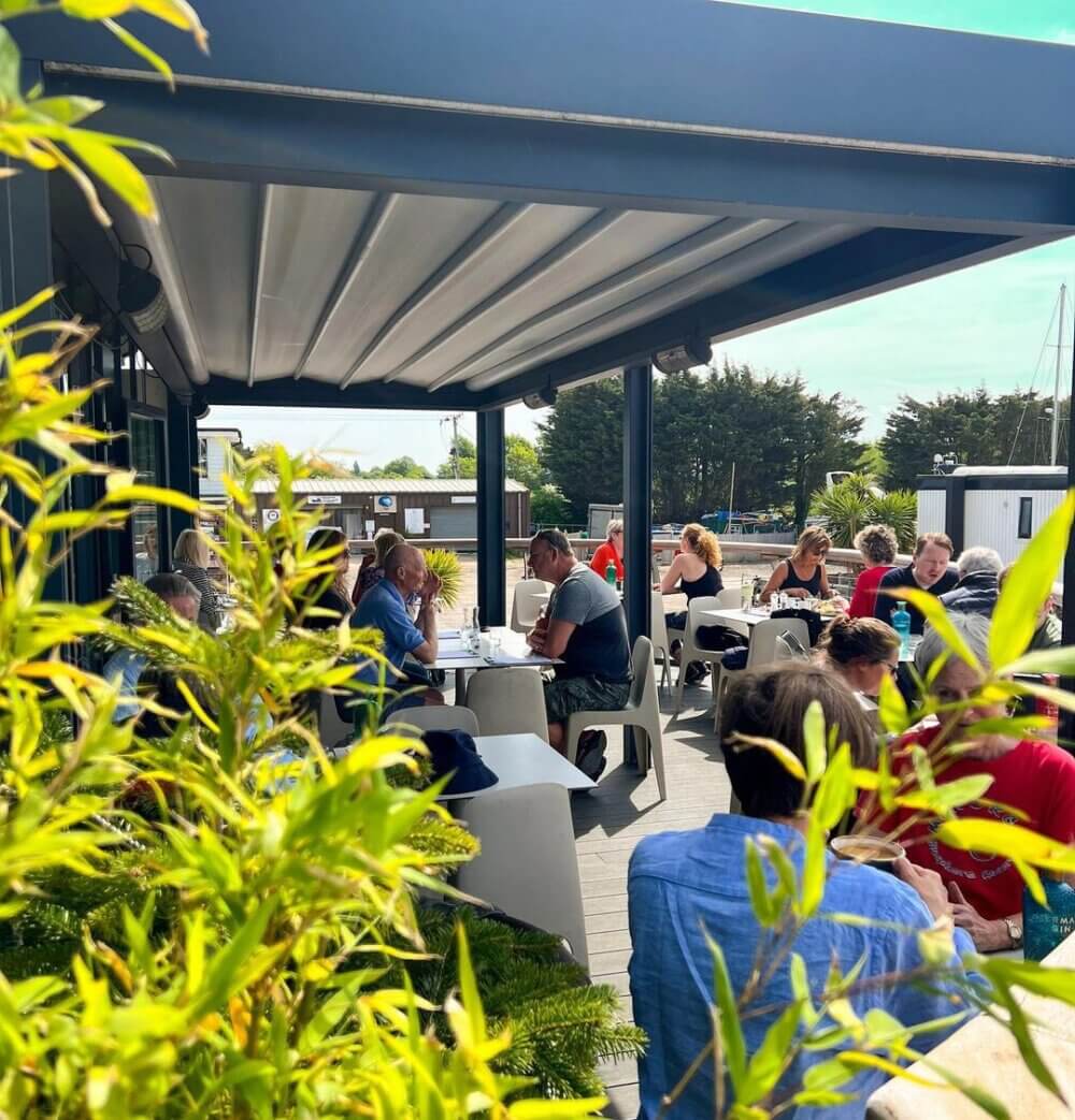 People dining on an outdoor patio under a shaded structure beside green plants with blue skies in the background. - Home Instead