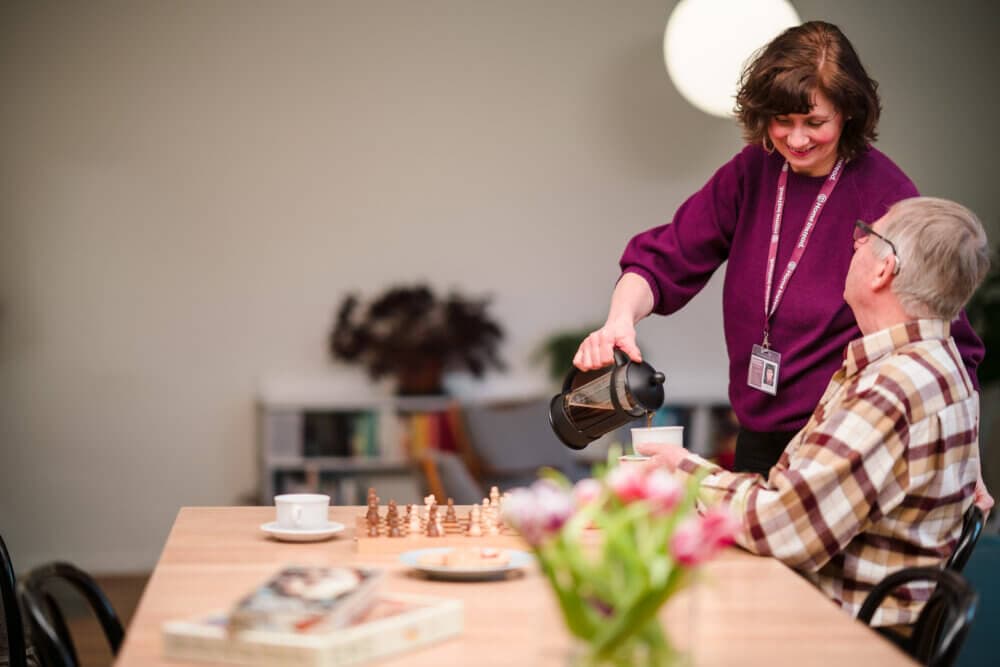 A woman pours coffee for a seated elderly man in a cozy room with a chessboard and books on the table. - Home Instead