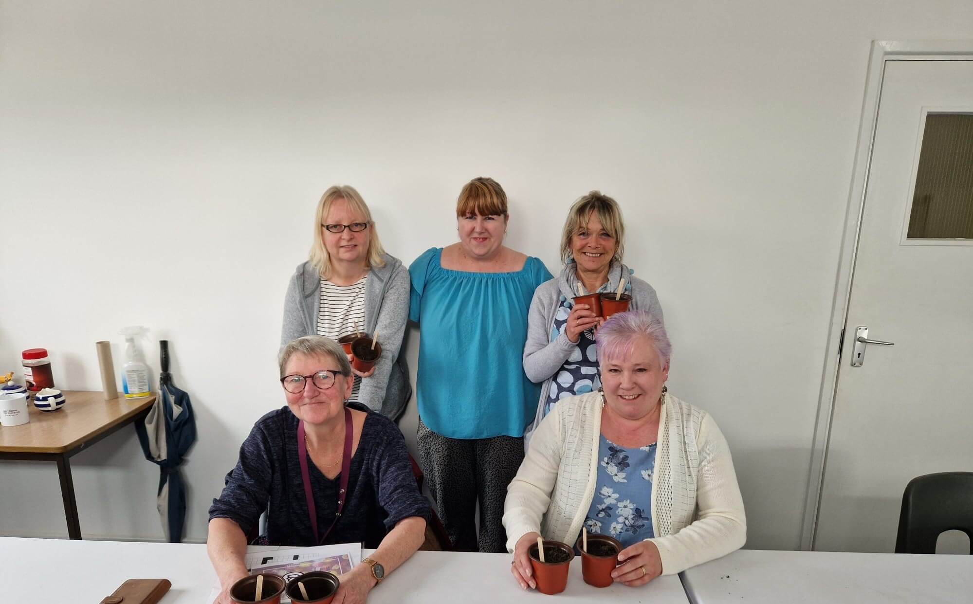 Five women, some holding small plant pots, smile while sitting and standing around a table in a white room. - Home Instead