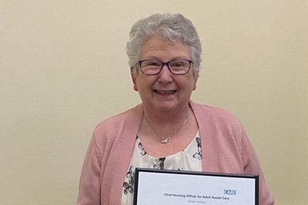 Smiling elderly woman with glasses holding an award in a frame, wearing a pink cardigan over a floral blouse. - Home Instead