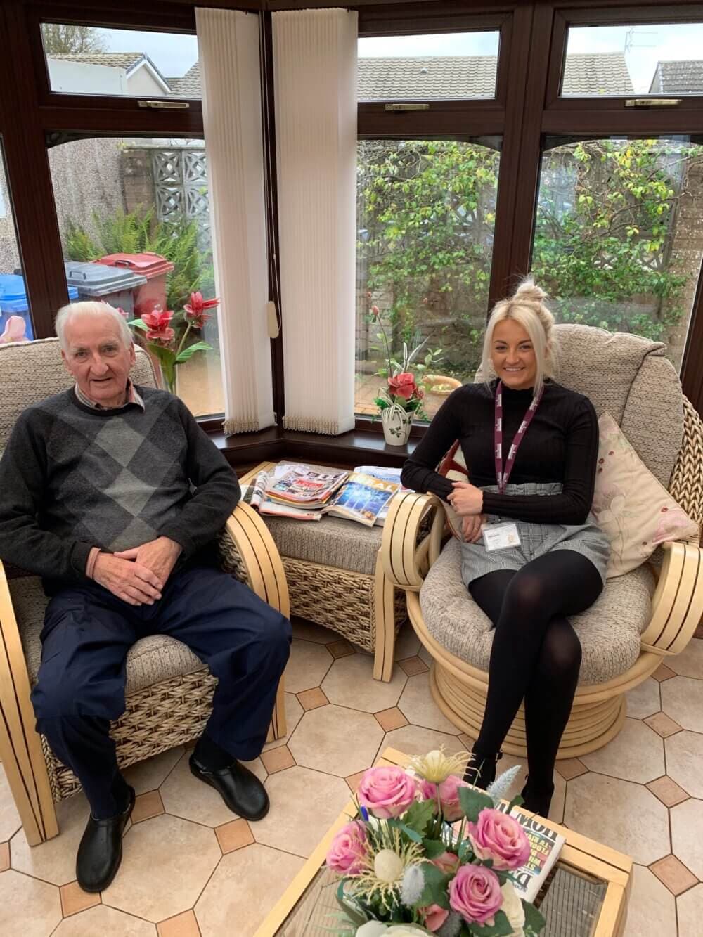 An elderly man and a young woman sit in a sunlit room with large windows, surrounded by flowers and magazines. - Home Instead