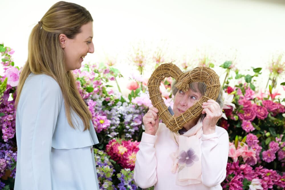 Two women sharing a moment, one holding a heart-shaped wicker frame, surrounded by colorful flowers. - Home Instead