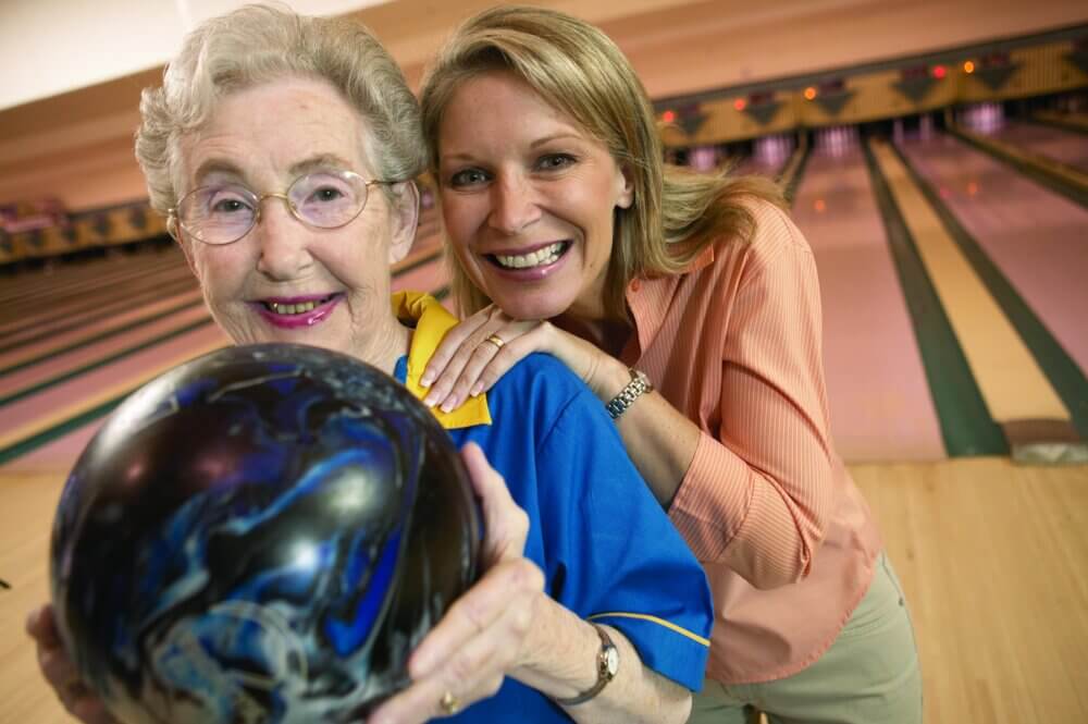 Older woman holding a bowling ball with younger woman leaning on her shoulder, both smiling at a bowling alley. - Home Instead
