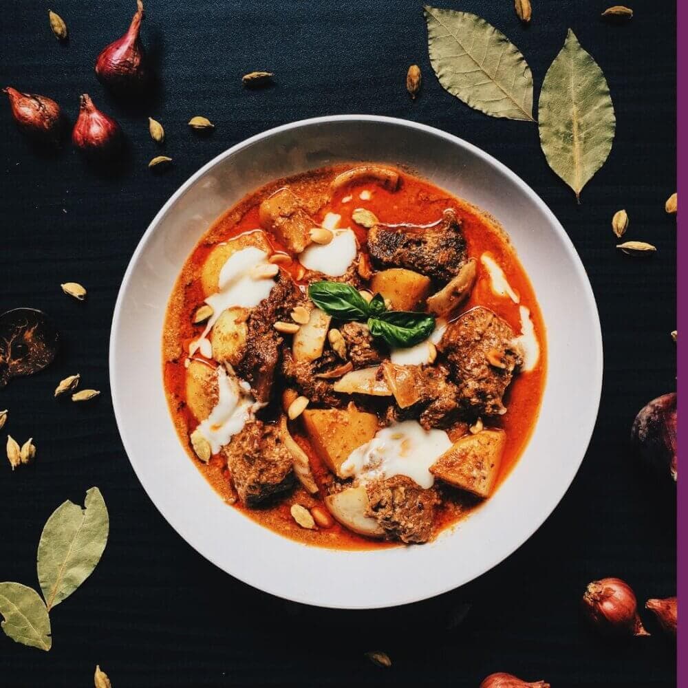 A bowl of spicy curry with meat and potatoes, garnished with leaves, surrounded by shallots and bay leaves on a dark table. - Home Instead