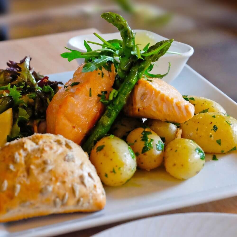 A plate of baked salmon, asparagus, baby potatoes, mixed greens, and a piece of bread on a white dish. - Home Instead