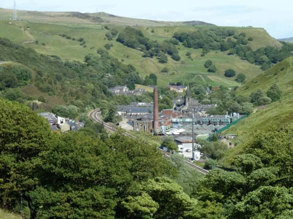 Aerial view of a small village with industrial buildings, surrounded by green hills and trees, with a railway passing through. - Home Instead