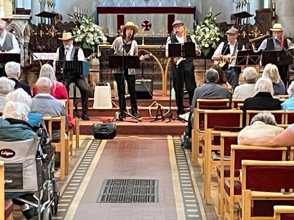 A music band performs in a church for an audience; some people sit, while others are in wheelchairs. - Home Instead