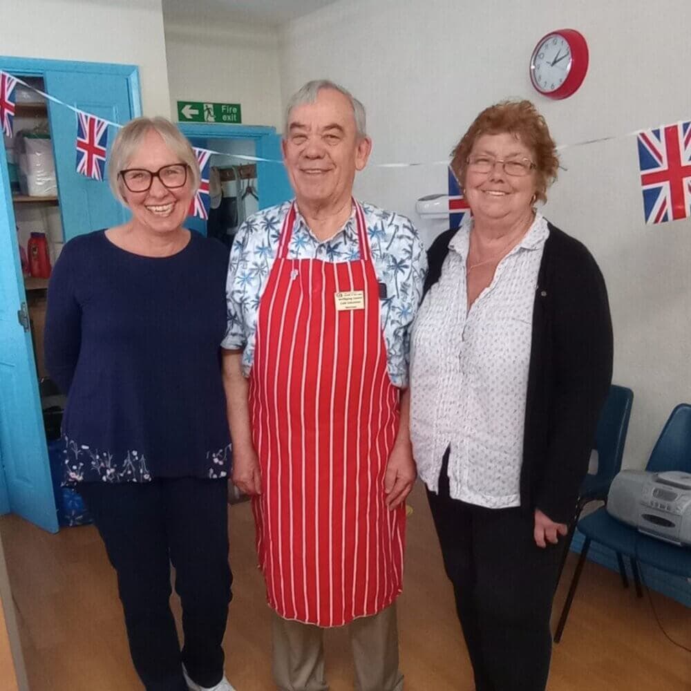 Three people smiling, one wearing a red-striped apron, standing in a room with Union Jack bunting and a clock on the wall. - Home Instead