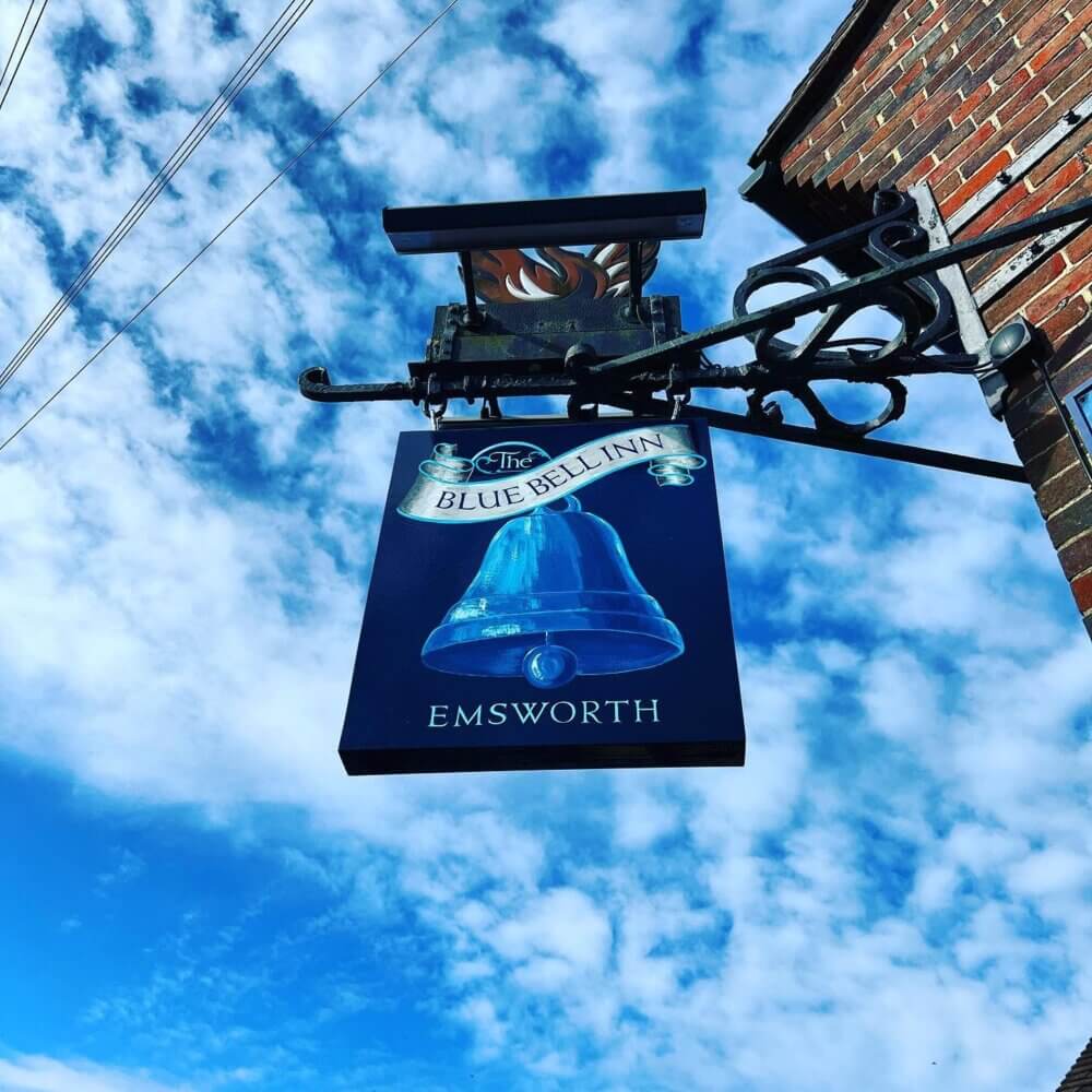 A pub sign for "The Blue Bell Inn" in Emsworth, featuring a blue bell, against a blue sky with scattered clouds. - Home Instead