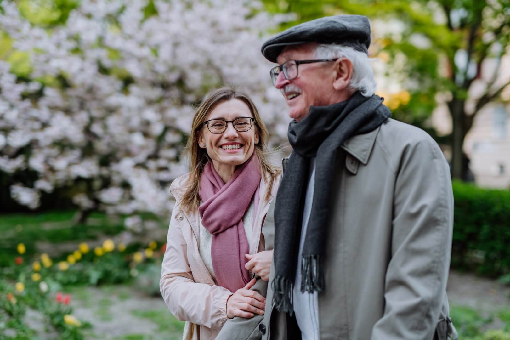 A smiling woman and an elderly man with glasses and a hat walk arm-in-arm in a garden with blooming trees. - Home Instead