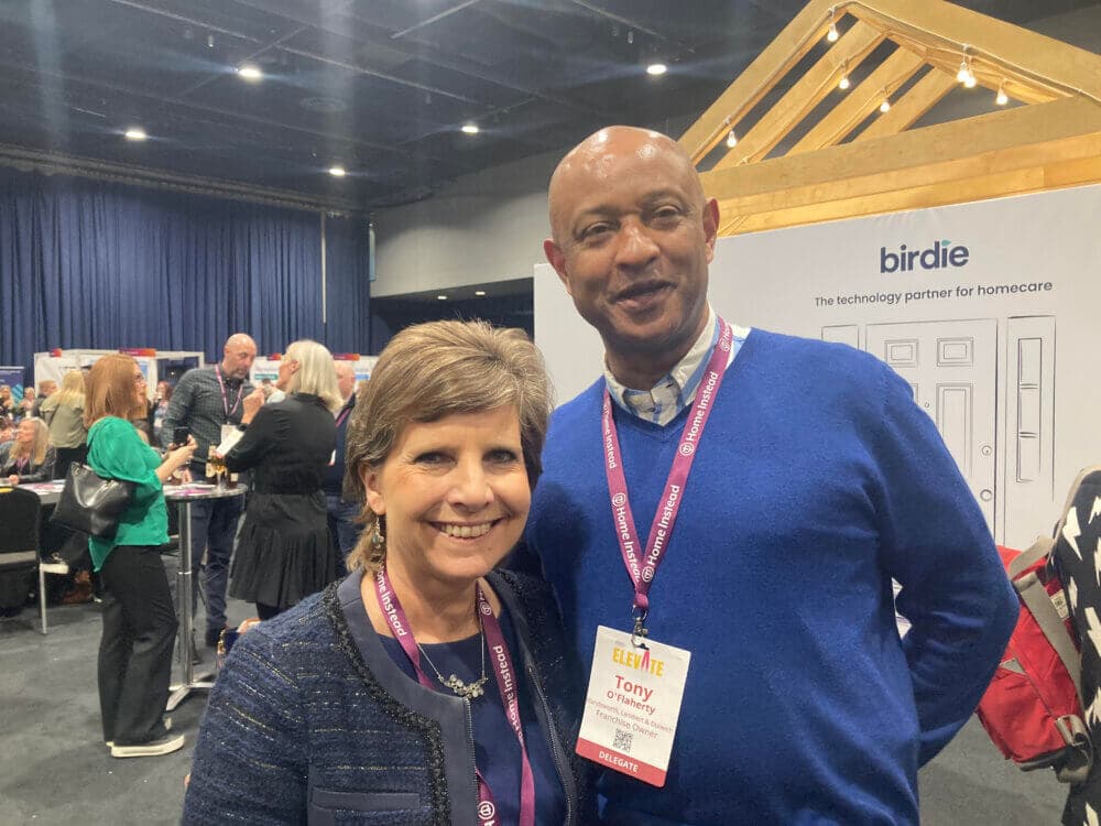 A smiling man and woman with visitor badges, standing in an event space with people and an exhibit in the background. - Home Instead
