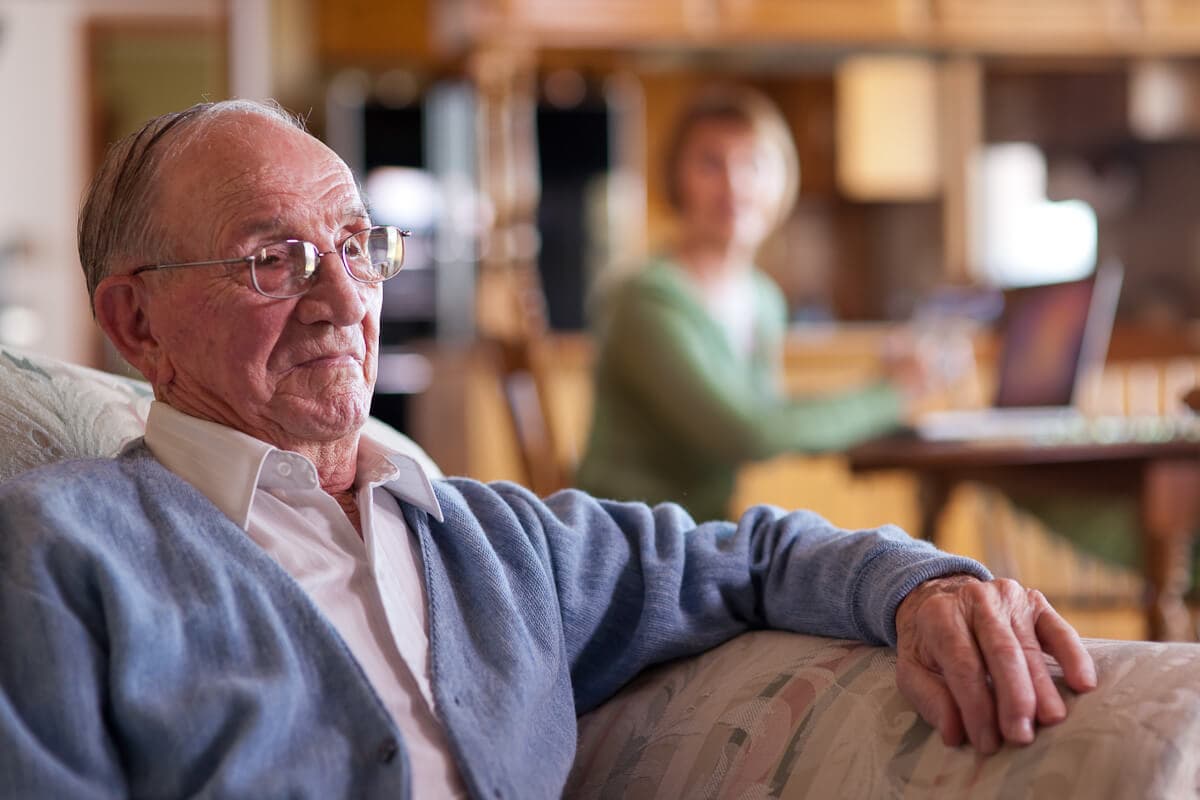 Elderly man sitting on a couch, looking thoughtful, with a woman working on a laptop in the background. - Home Instead