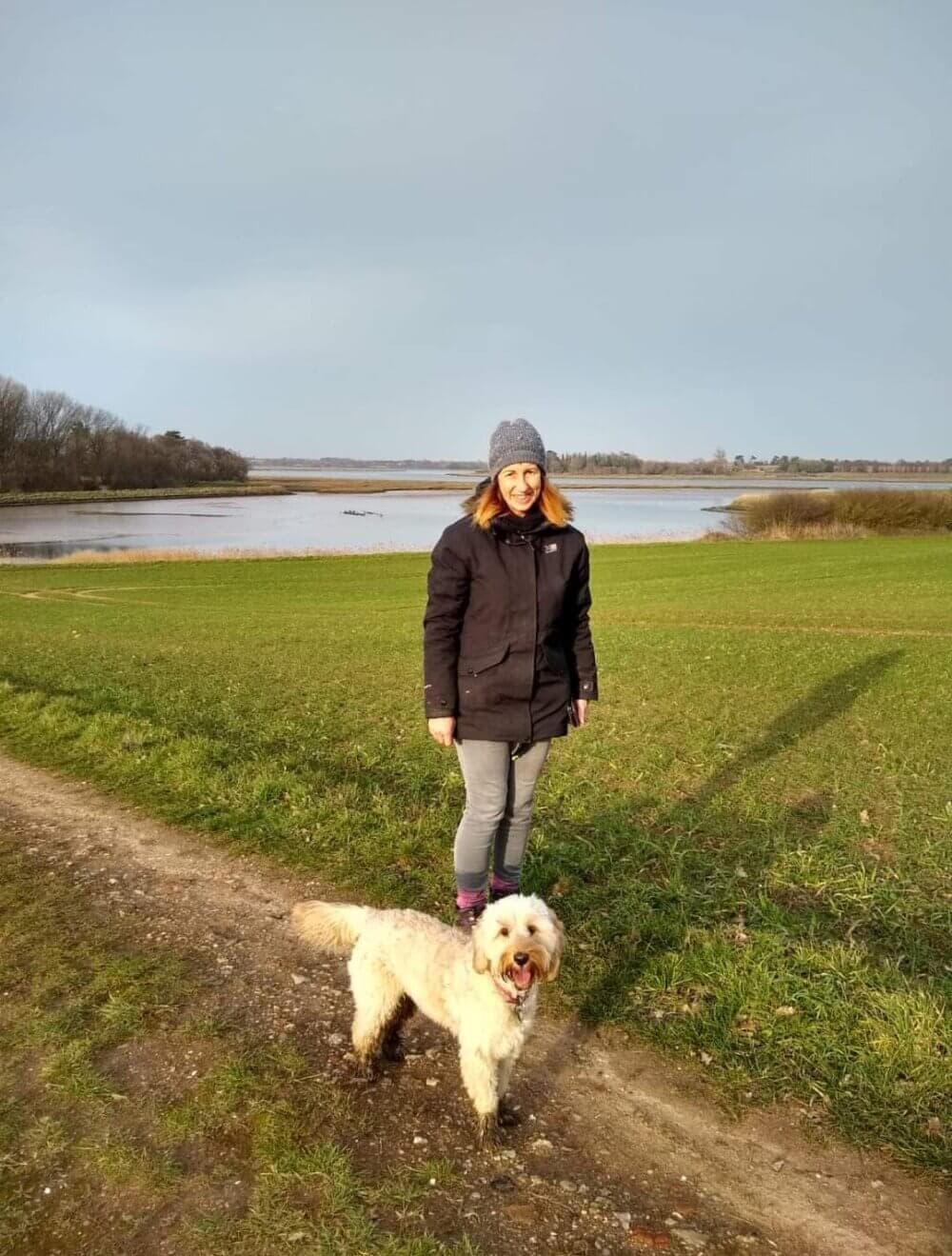 Person in a jacket and hat stands on a grassy path near a large body of water, with a fluffy dog beside them. - Home Instead