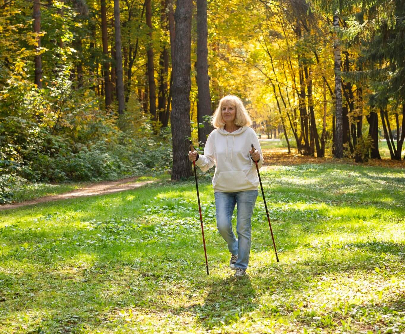 A person with blonde hair in a white hoodie walks with hiking poles on a grassy path in a sunlit forest. - Home Instead