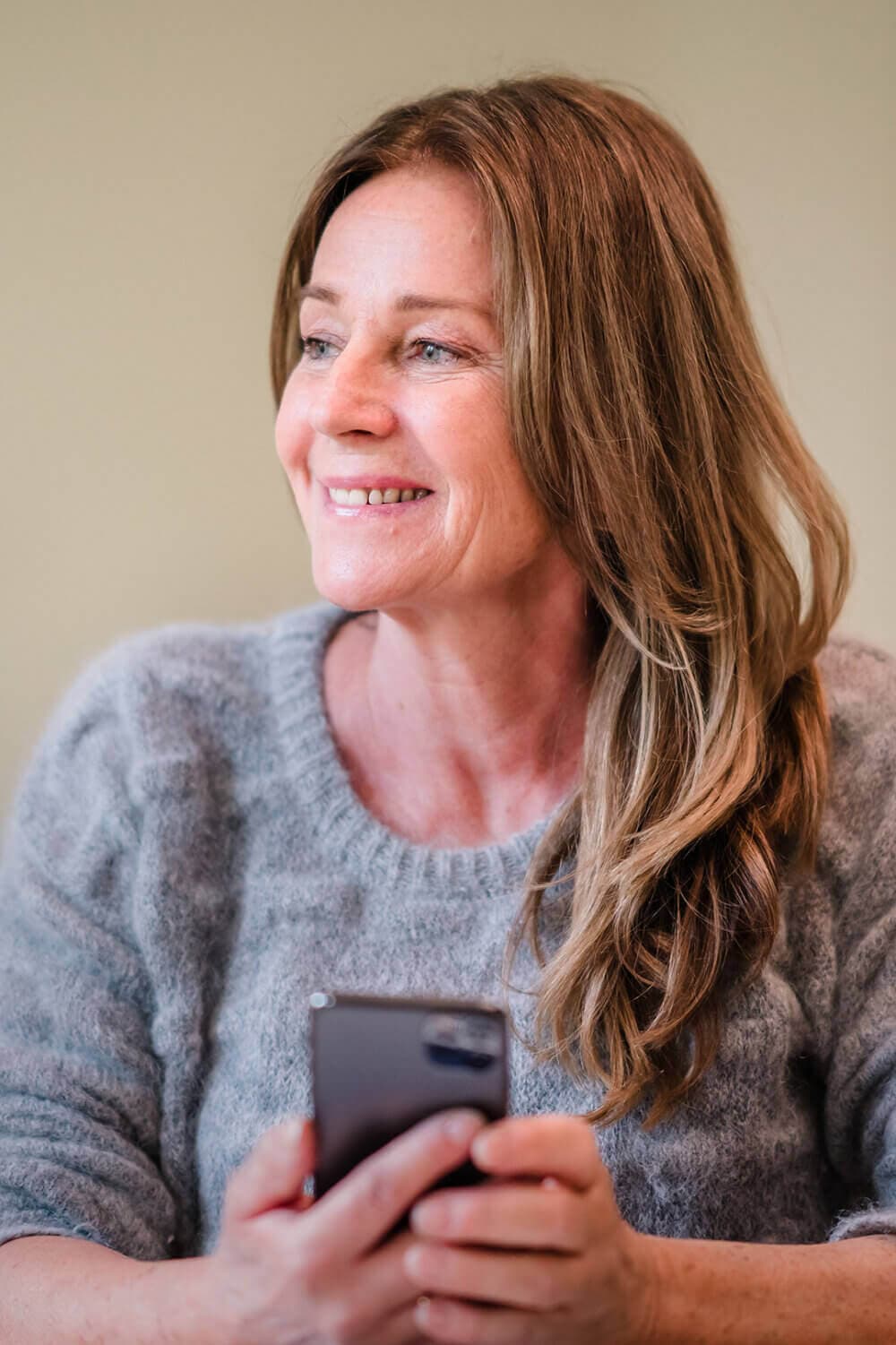 Woman smiling while holding a smartphone, wearing a gray sweater, sitting against a light background. - Home Instead