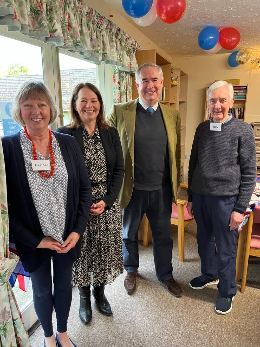 Four people stand in a room decorated with balloons and streamers, wearing name tags, smiling at the camera. - Home Instead