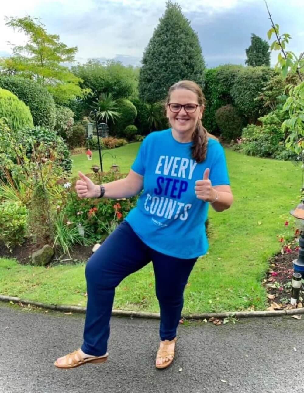 Person in blue "Every Step Counts" shirt giving thumbs-up, standing on a garden path surrounded by greenery. - Home Instead