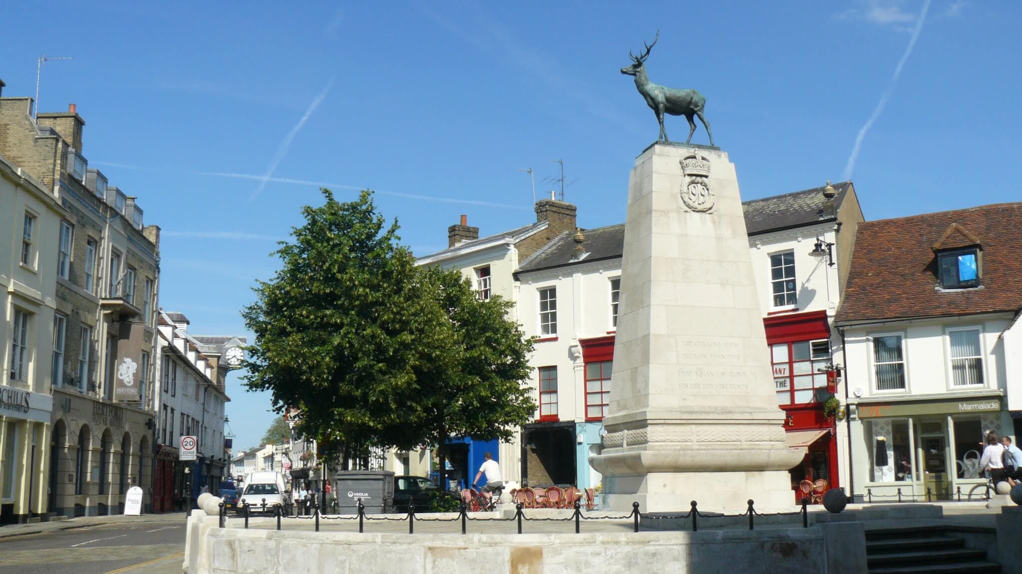 A town square with a monument featuring a stag statue atop, surrounded by buildings and a tree. - Home Instead