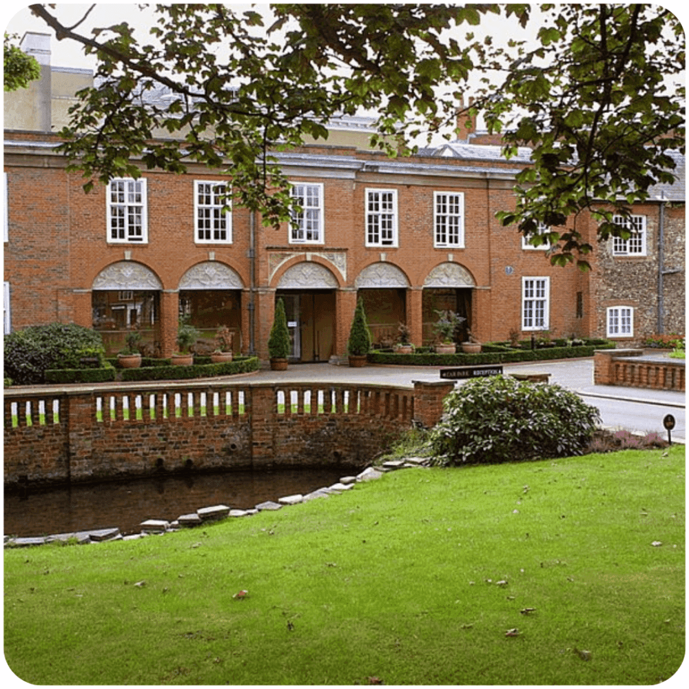 Brick building with arched windows, a pond and greenery in the foreground, framed by tree branches. - Home Instead