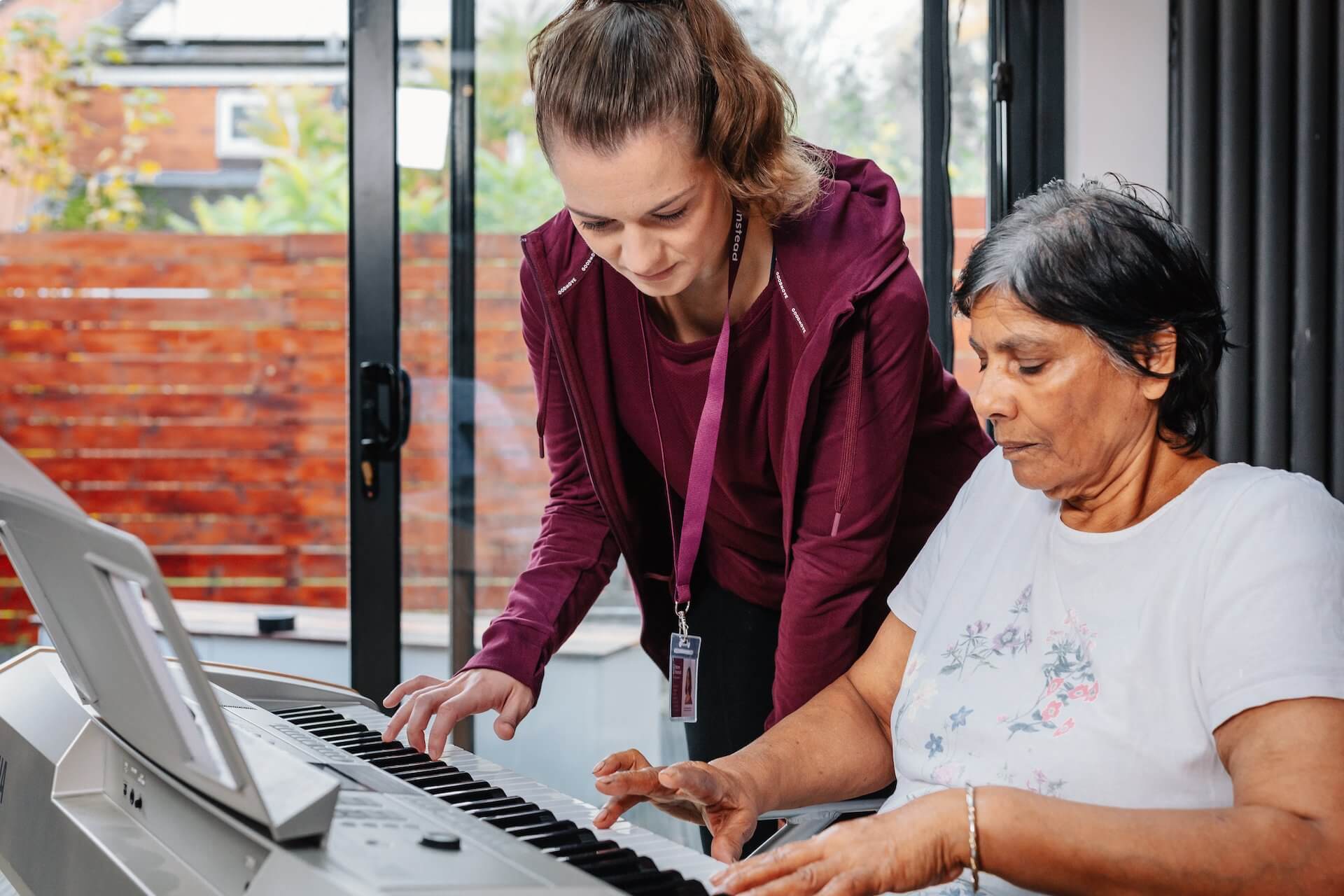 A senior woman playing the piano with assistance from a younger woman standing beside her indoors. - Home Instead