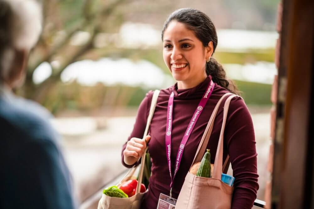 A woman with bags of groceries talks to someone at a doorway, smiling, and wearing an ID badge. - Home Instead