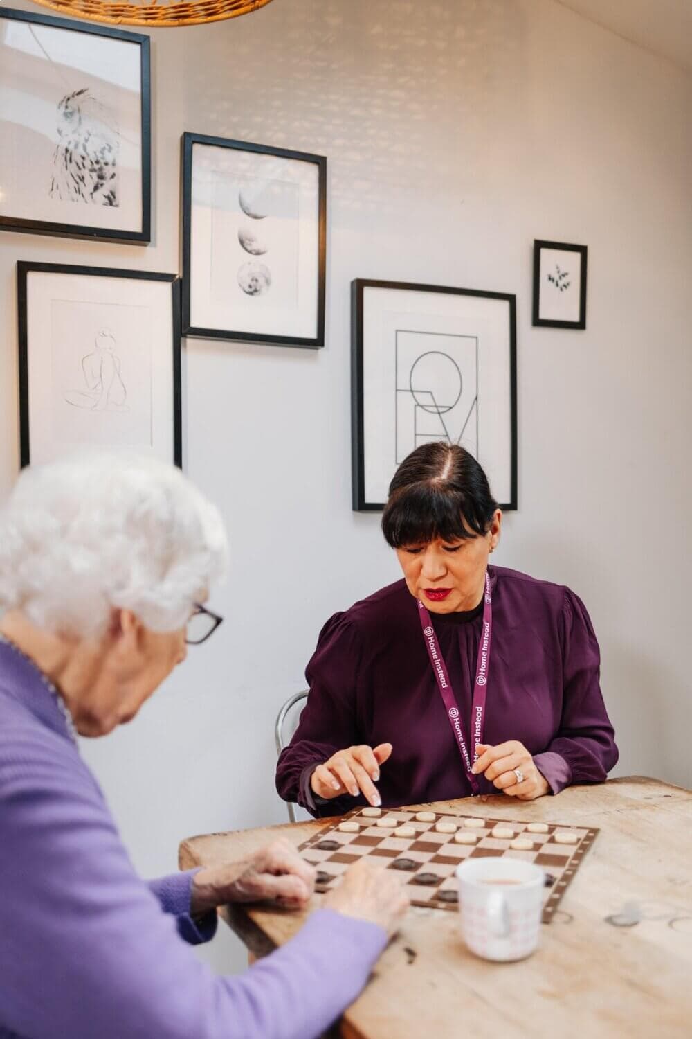 Two women are playing a board game at a wooden table, with framed art on the wall behind them. - Home Instead