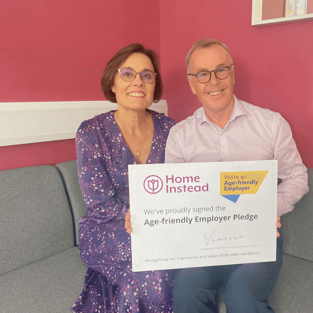 Two people sitting on a couch, holding a sign announcing Home Instead's Age-friendly Employer Pledge in a pink room. - Home Instead