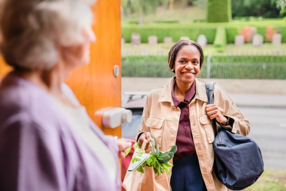 A woman holding groceries smiles at another woman standing in a doorway, with a garden blurred in the background. - Home Instead