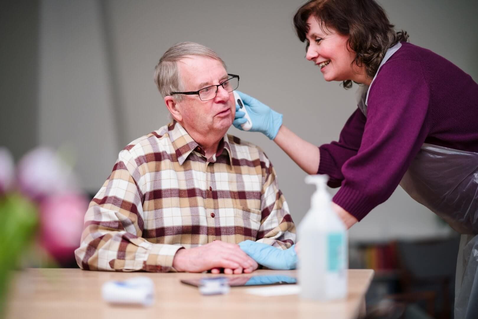 A woman in gloves gently wipes an older man's face while he sits at a table, with hygiene items nearby. - Home Instead