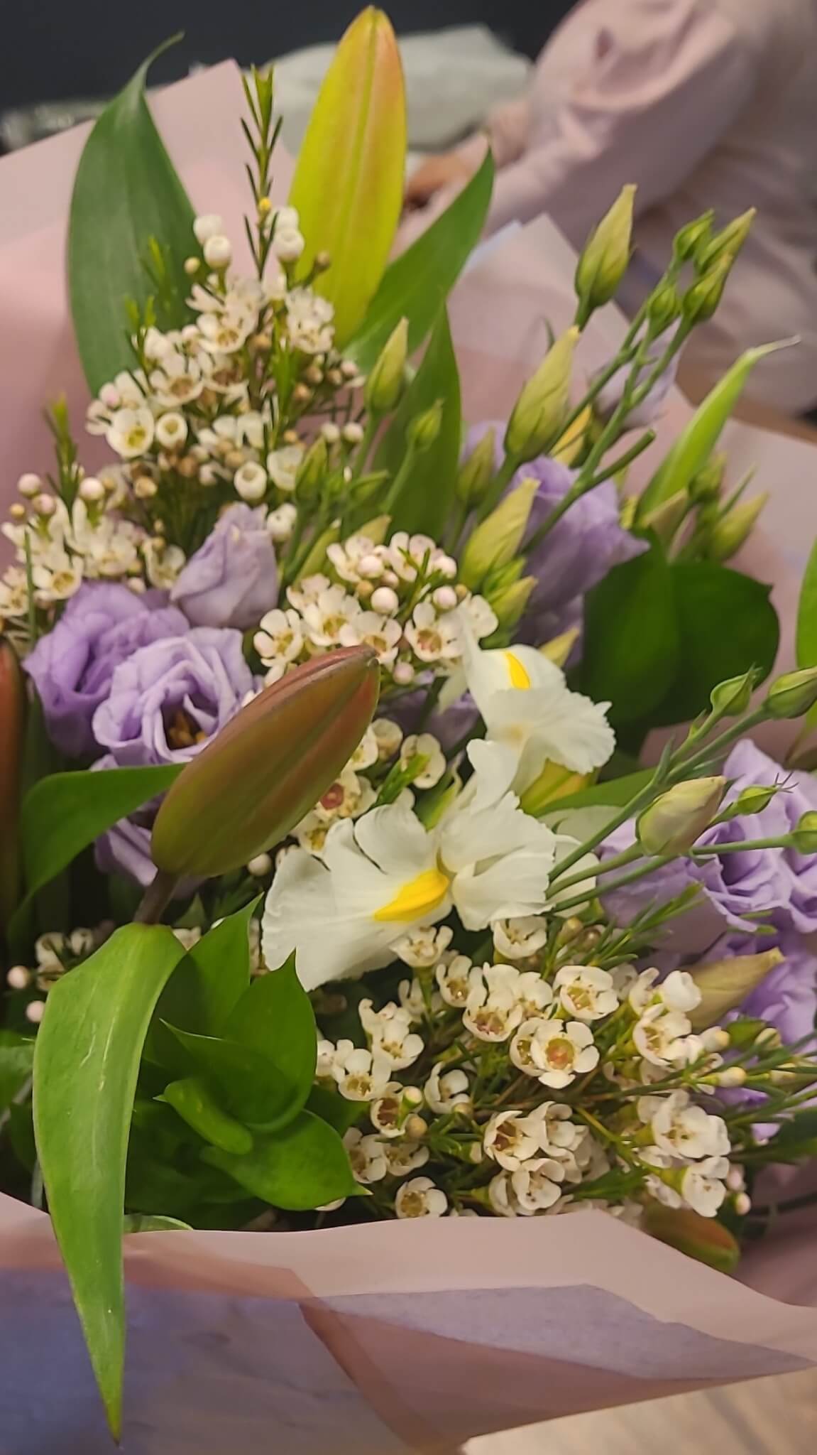 A bouquet with purple roses, white lilies, and small white flowers, wrapped in pink paper. - Home Instead