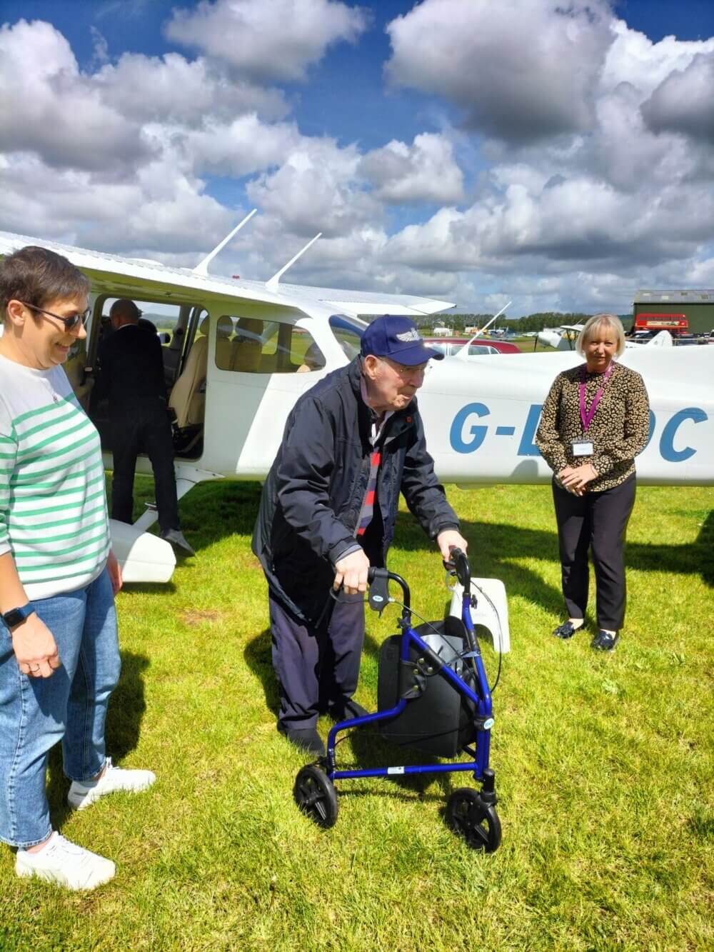An elderly man using a walker is assisted as he exits a small airplane, with two women standing nearby on a grassy field. - Home Instead