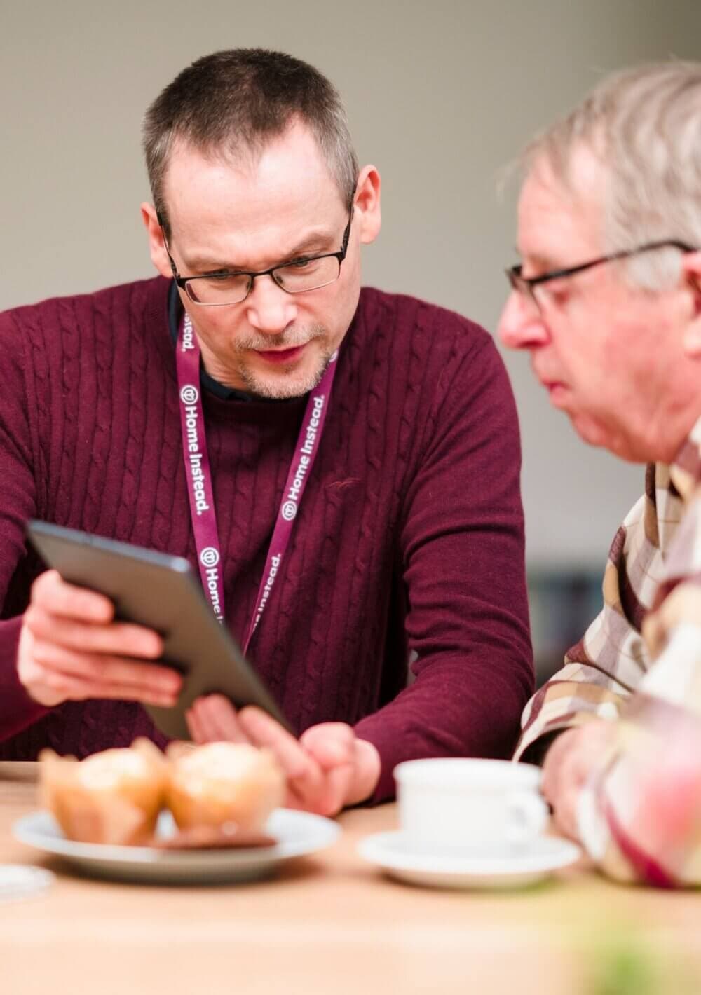 Two men sitting at a table, one holding a tablet, with coffee cups and muffins in front of them. - Home Instead
