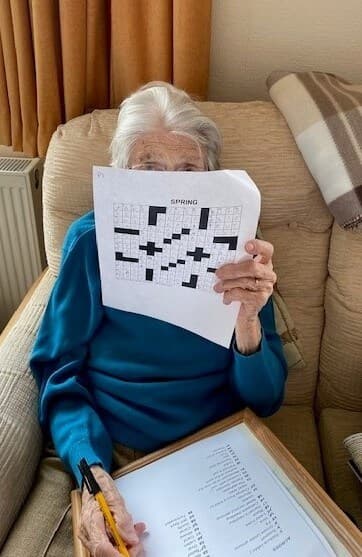 An elderly person holds up a crossword puzzle while sitting on a beige couch, their face partially obscured. - Home Instead
