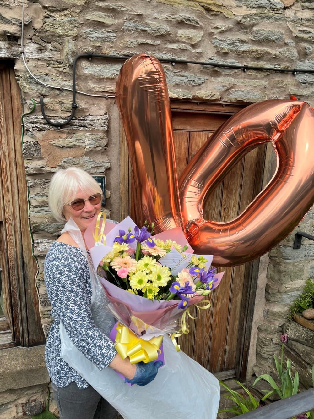 A woman holding a large bouquet of flowers and a balloon shaped like the number 70, standing in front of a stone wall. - Home Instead