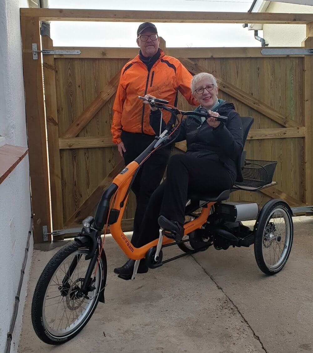 Two people in front of a wooden gate. One stands while the other sits on an orange recumbent tricycle, both smiling. - Home Instead