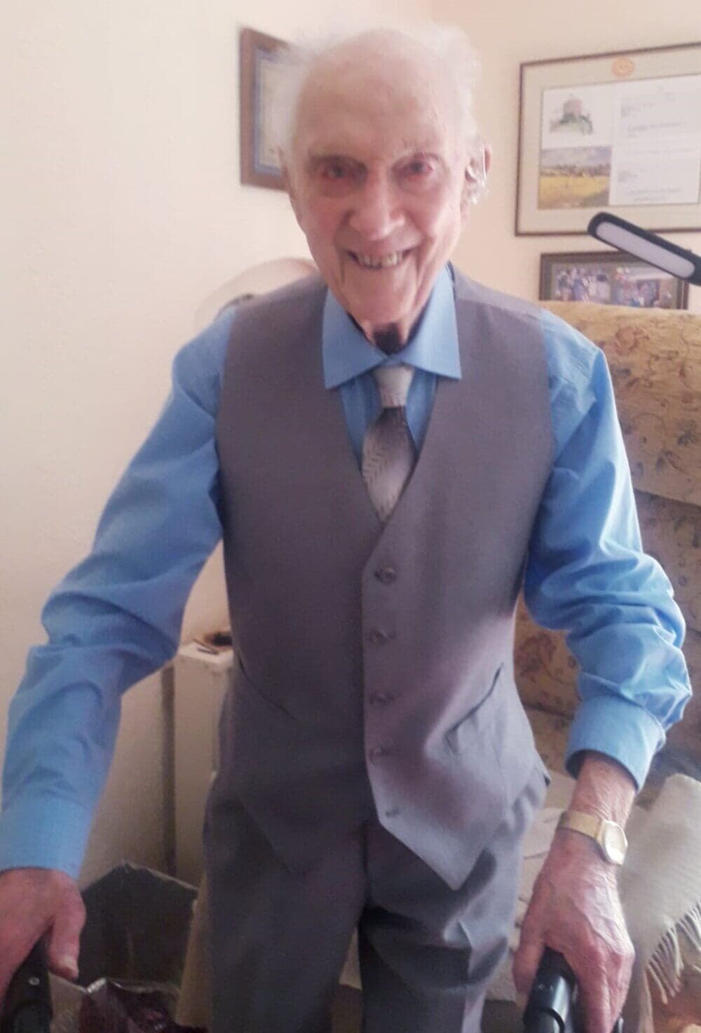An elderly man in a blue shirt and gray vest smiles while using a walker in a warmly decorated room. - Home Instead