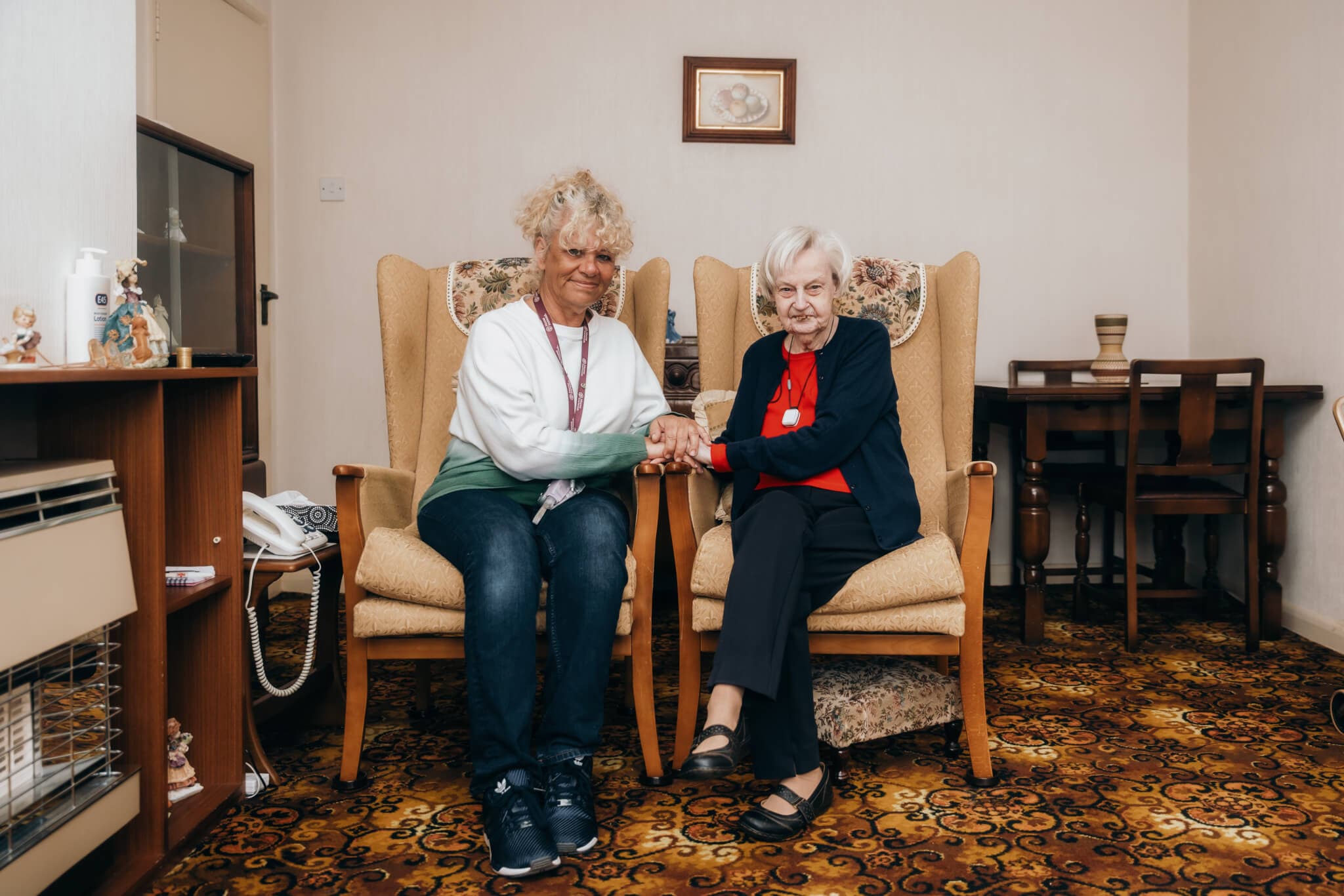 Two elderly women sitting on armchairs, holding hands and smiling inside a cozy, carpeted living room. - Home Instead