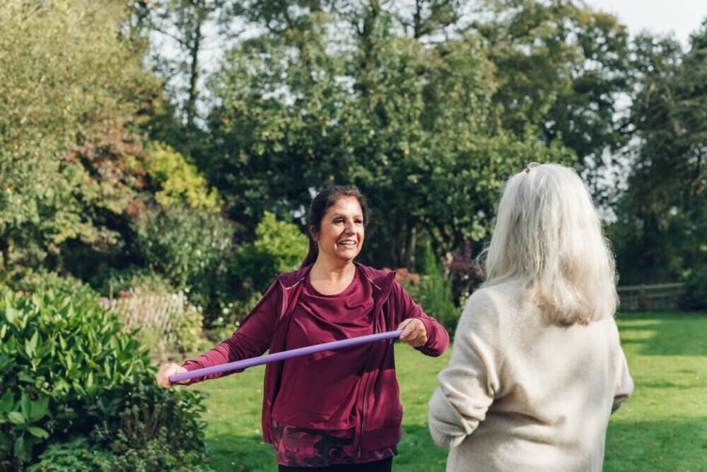 Two women exercising with a resistance band outdoors in a leafy park, one pulling the band and smiling at the other. - Home Instead