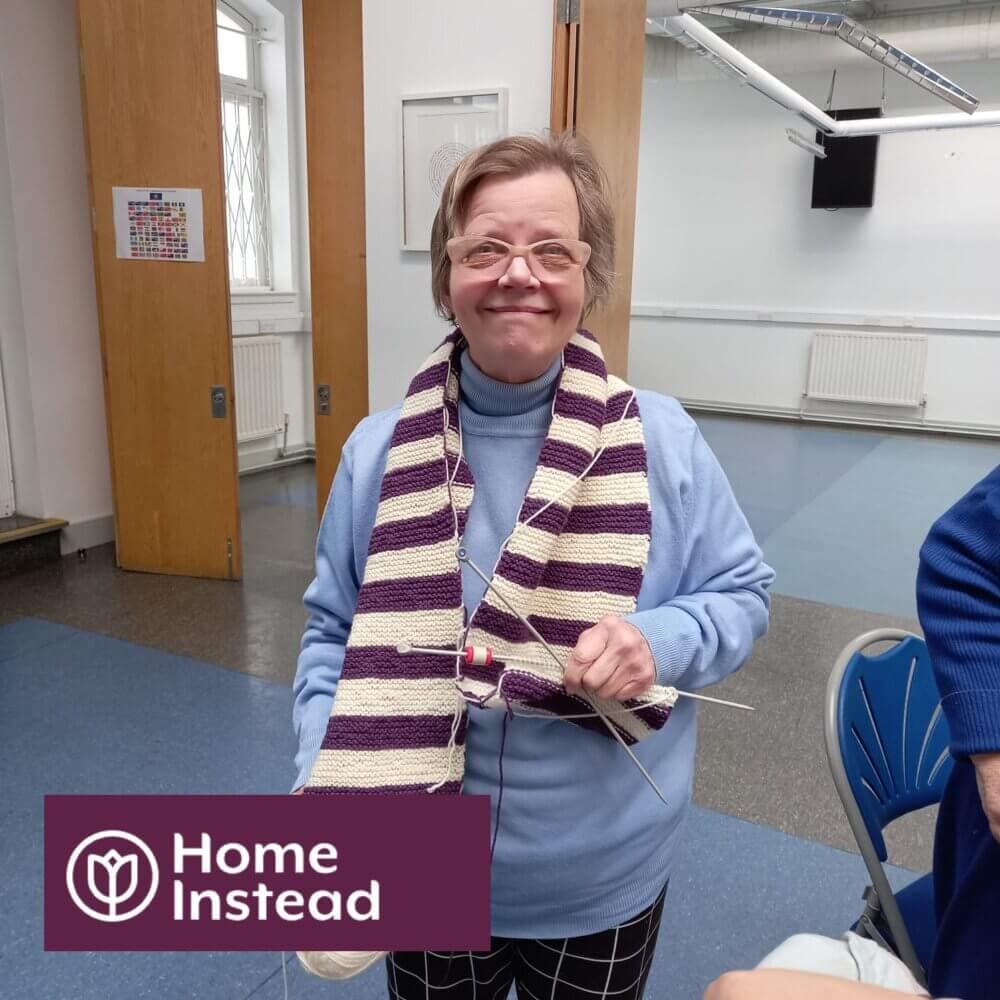 Elderly woman smiling and knitting with yarn in a room, wearing a striped scarf. "Home Instead" logo in bottom left. - Home Instead