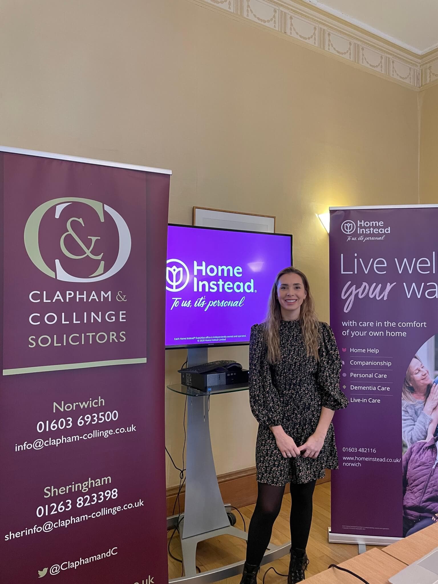 A woman stands between banners for Clapham & Collinge Solicitors and Home Instead Senior Care in an office setting. - Home Instead