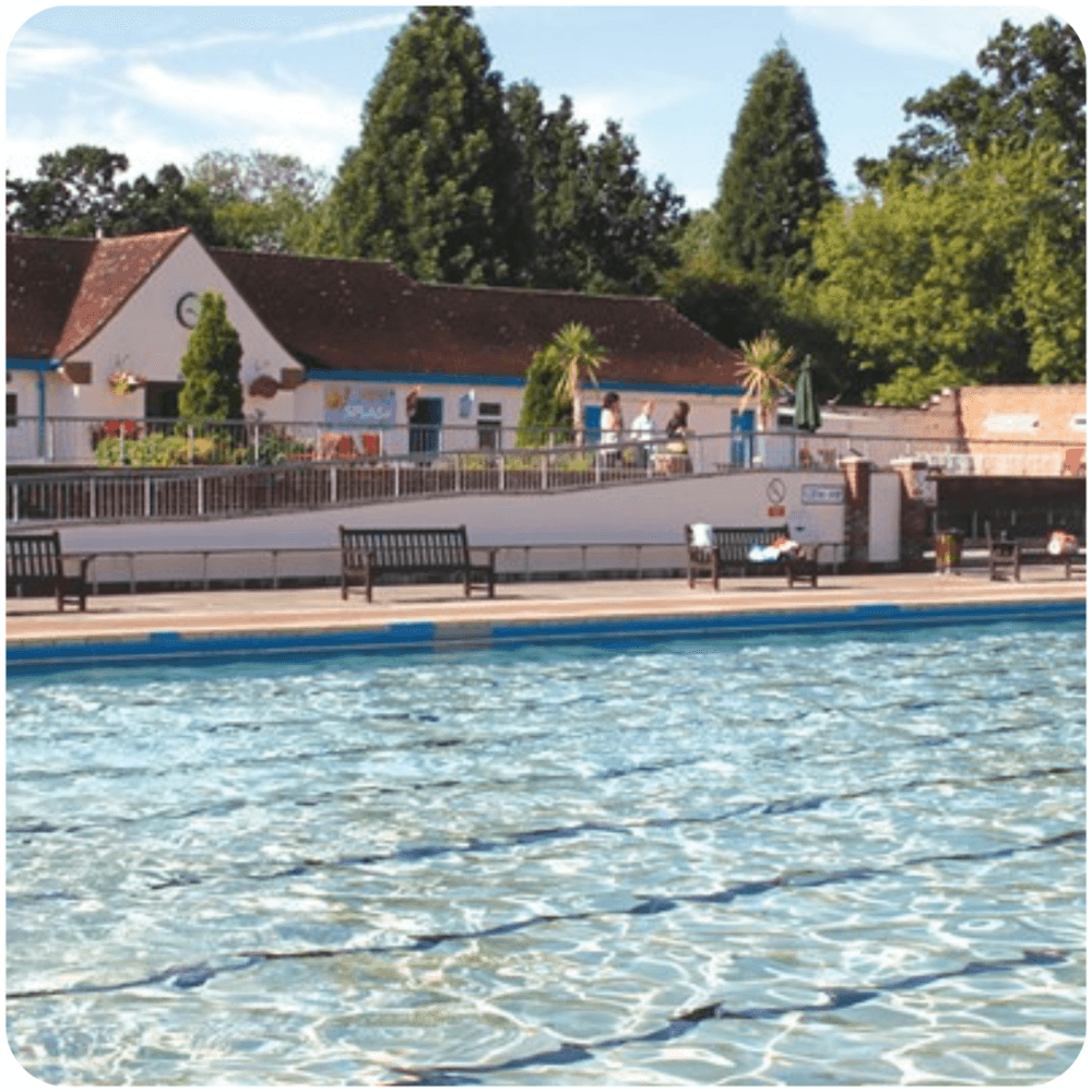 An outdoor pool with benches, surrounded by trees and buildings under a clear, blue sky. - Home Instead