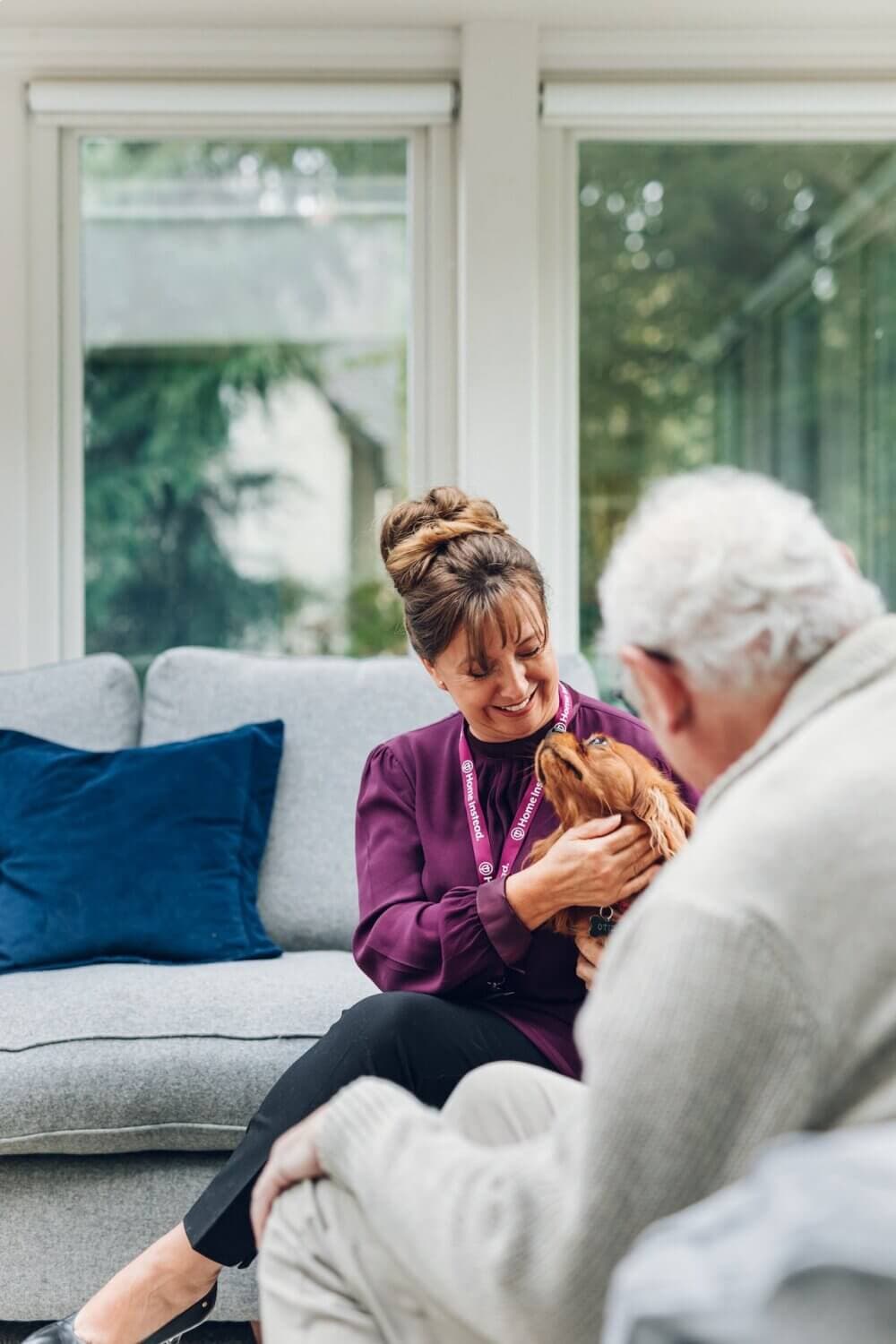 A woman in a purple shirt and elderly man with white hair smile at a small dog in a bright living room. - Home Instead Southampton