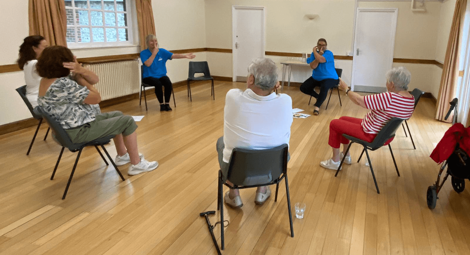 A group of elderly people participating in a seated exercise class in a bright room with wooden floors and large windows. - Home Instead