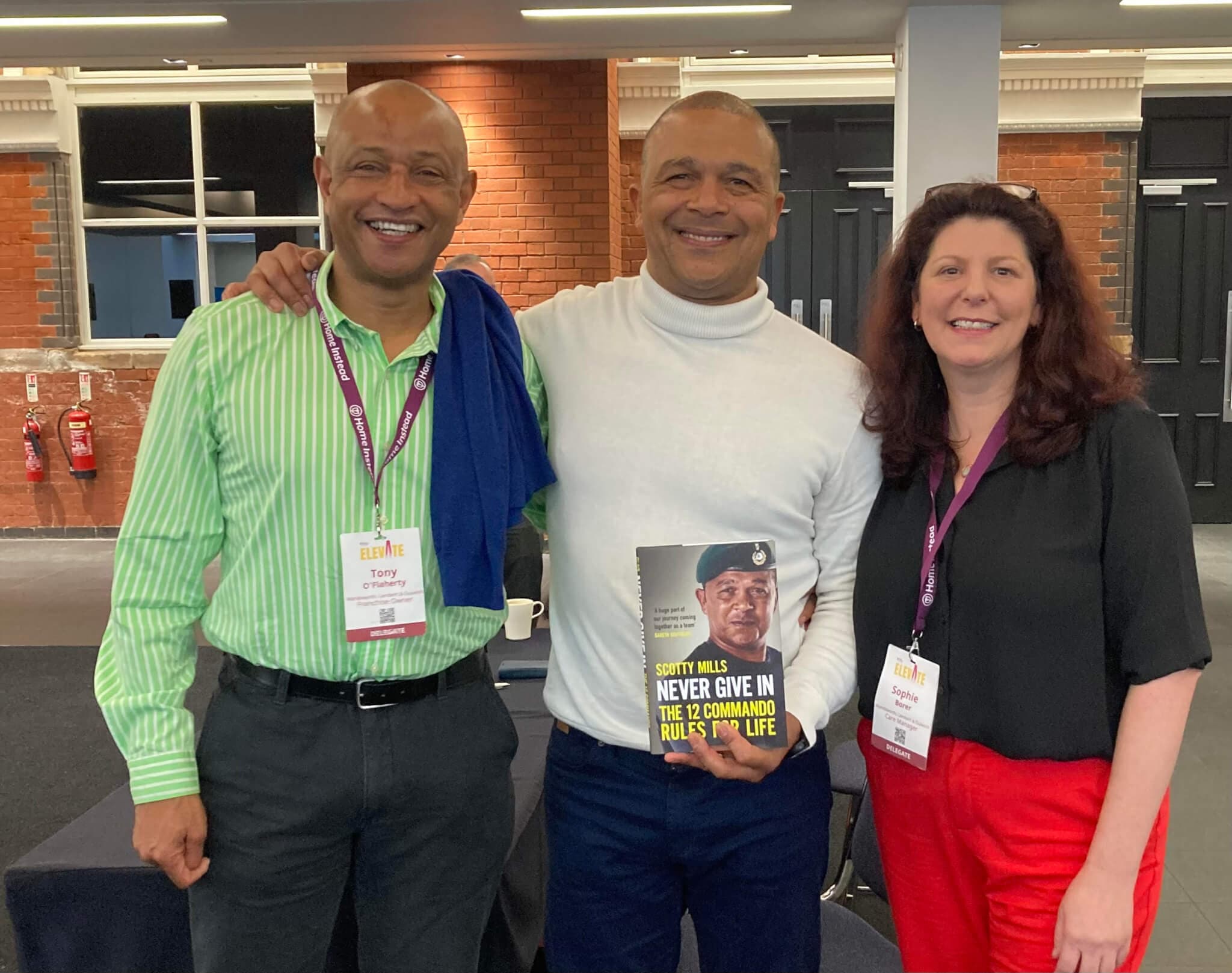 Three smiling people stand together; the middle person holds a book. They all wear conference badges. - Home Instead