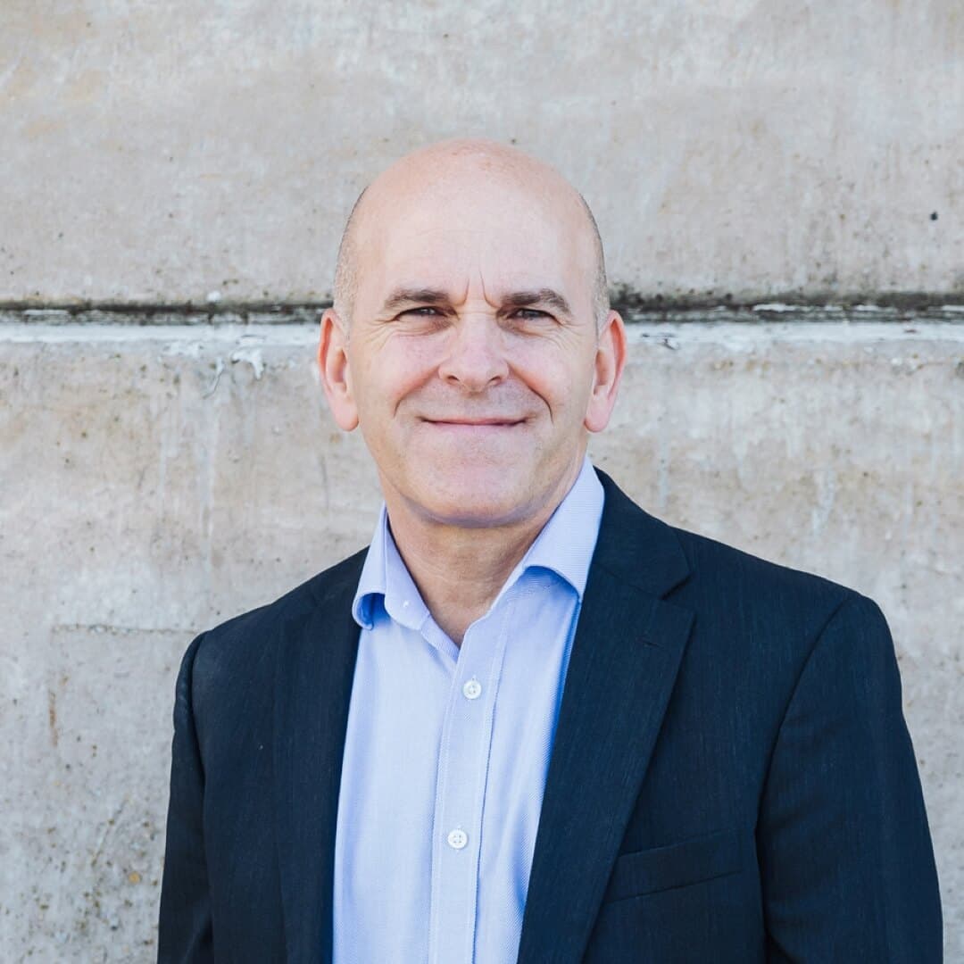 A bald man in a dark suit and light shirt smiles in front of a textured concrete wall. - Home Instead