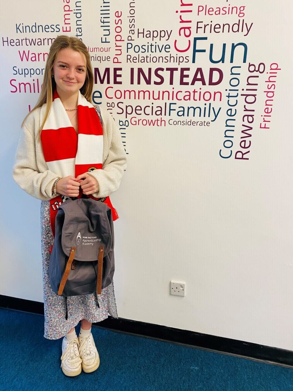 Young woman smiling, wearing a red and white scarf, holding a backpack, standing in front of a word-filled wall background. - Home Instead